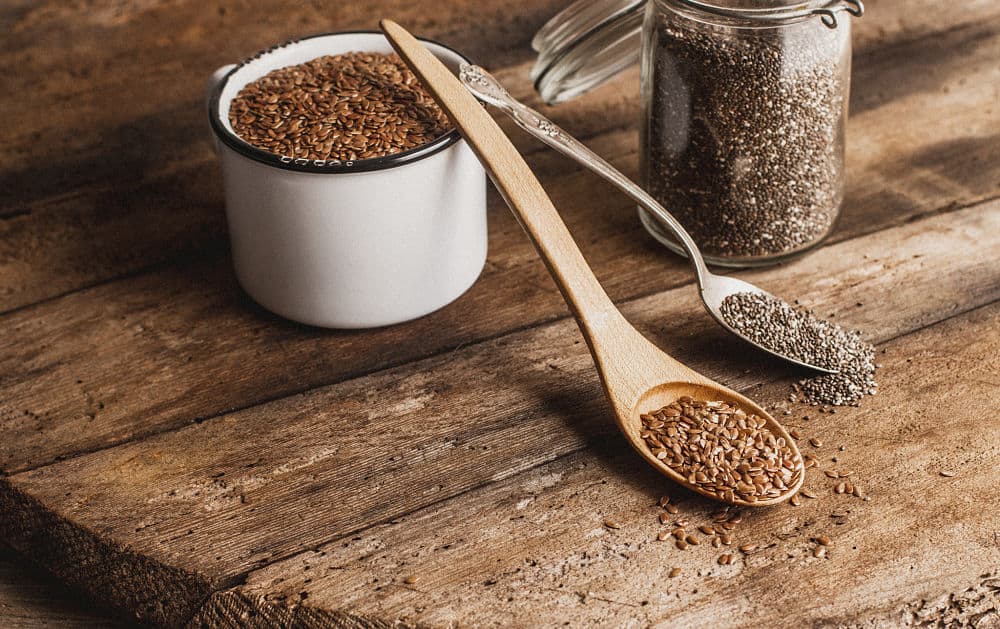 Flax seeds in a white enamal cup and chia seeds in a small Mason jar with spoonful of both grains leaning on the white cup