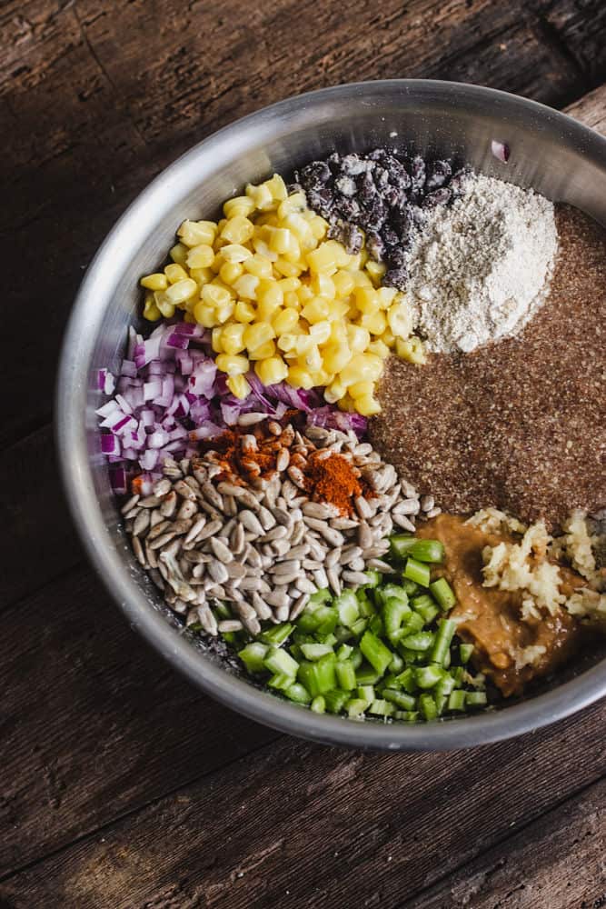 Ingredients in sections in a bowl: smashed cooked blacks beans, corn, diced red onions, Cayenne pepper on top of sunflower seeds, chopped celery, crushed garlic on top of miso loose paste, ground meal and oar flour