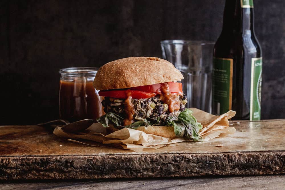 A burger on a narrow wooden board with ketchup oozing our of it in front of a small jar of bbq sauce, an empty glass and a beer