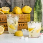 Two glasses of lemonade in front of a small wood container of lemons