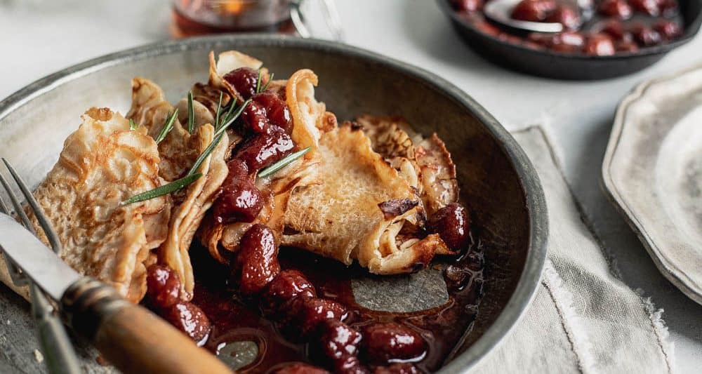 A close-up of folded pancakes in a metallic plate with strawberry sauce dripping with a small jar of maple syrup behind it and a small skillet of strawberry sauce on the side