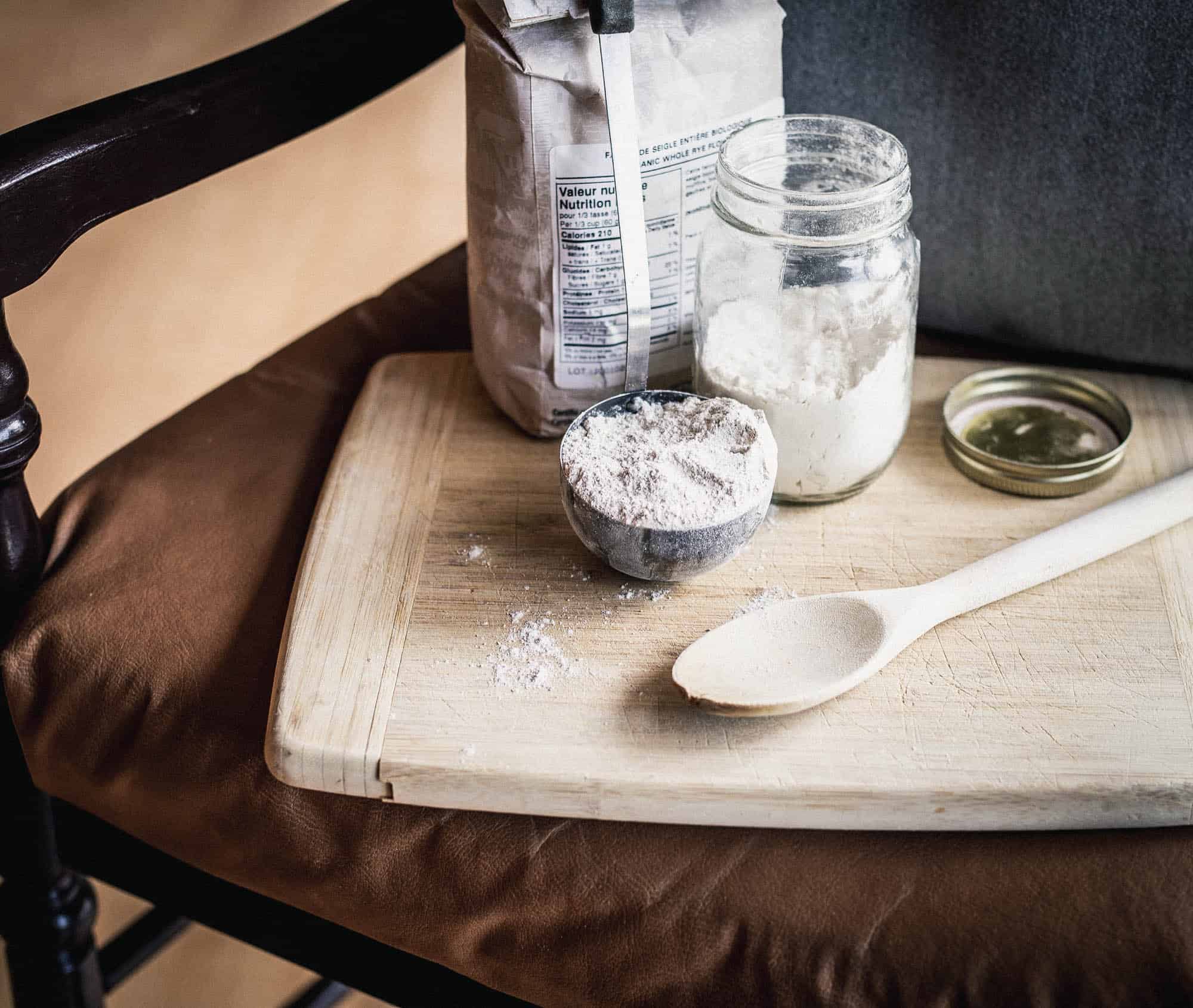 A bag of flour, a laddle of rhye flour, a jar of white flour, and a small wooden spoon are standing on a wooden cutting board which is on a chair.