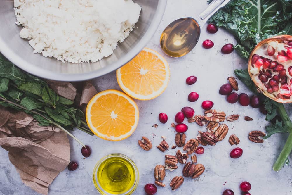 Top view of mint sprigs on brown paper, cauliflower rice in a mixing bowl, honey in a measuring spoon, kale leaf with half of a pomegranate, fresh cranberries and pecans spread apart, olive oil in a tiny mixing bowl, and an orange sliced in two