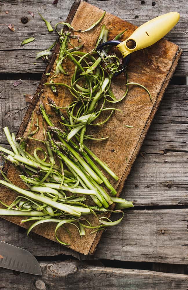 A bird's eye view of shaved asparagus in ribbons with a vegetable peeler on top of a narrow wooden cutting board 