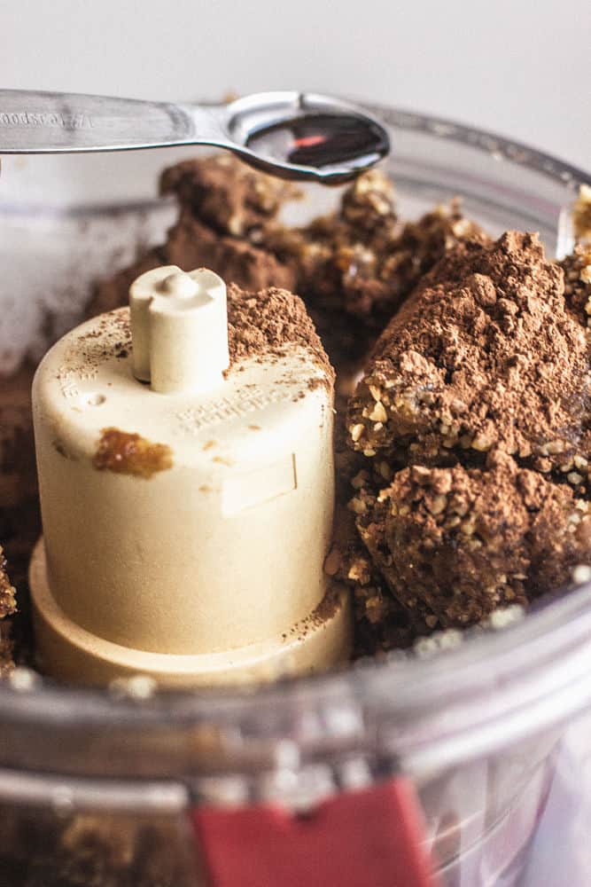 Close-up of brownie dough in a food processor bowl with a spoon of vanilla on the verge of being added