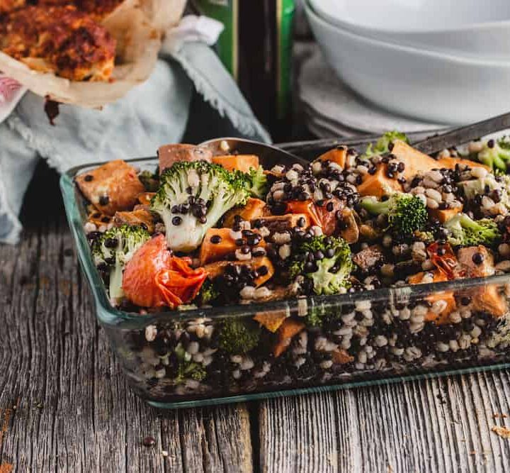 Lentil, barley, and sweet potato salad with broccoli and baby tomatoes in a glass baking dish sitting on a wooden board