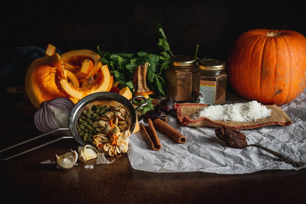 Nature morte (dans le sens d’une aiguille d’une montre ou de gauche à droite : citrouille coupée en deux, persil, deux petits pots de cannelle et de muscade, citrouille entière, flocons de noix de coco reposant sur un petit sac en jute, miso dans une cuillère, bâtons de cannelle, dattes Medjool, graines de citrouille vertes et jaunes dans un petit tamis, gousses d’ail, oignon rouge coupé en deux