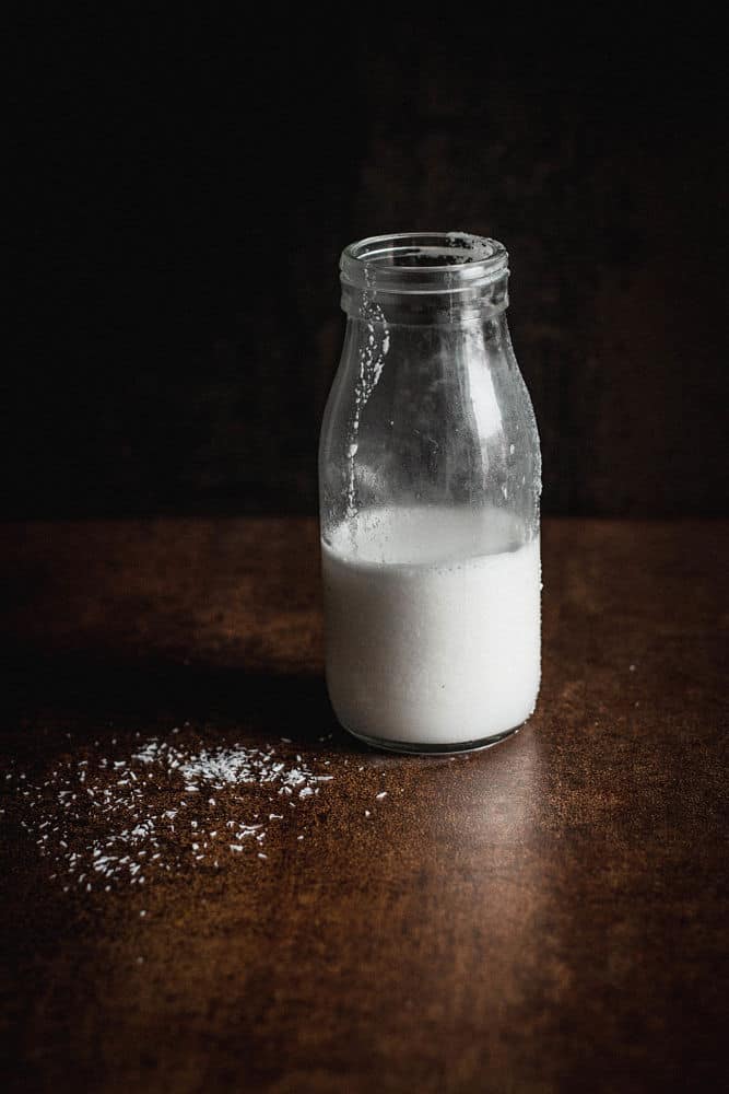 A small glass bottle with coconut milk at the right of spilled coconut flakes on a brown surface and a black background