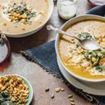 Overhead view of two bowls of pumpkin soup placed diagonally with a glass of red wine in the left corner and roasted pumpkin and squash seeds on a small plate