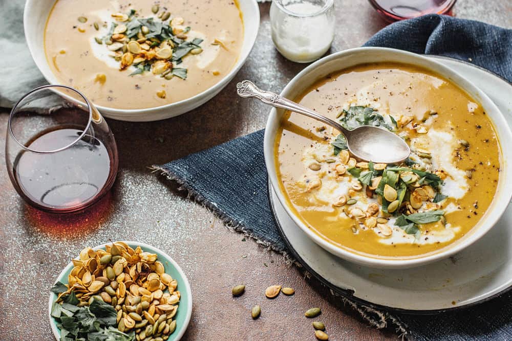 Overhead view of two bowls of pumpkin soup placed diagonally with a glass of red wine in the left corner and roasted pumpkin and squash seeds on a small plate