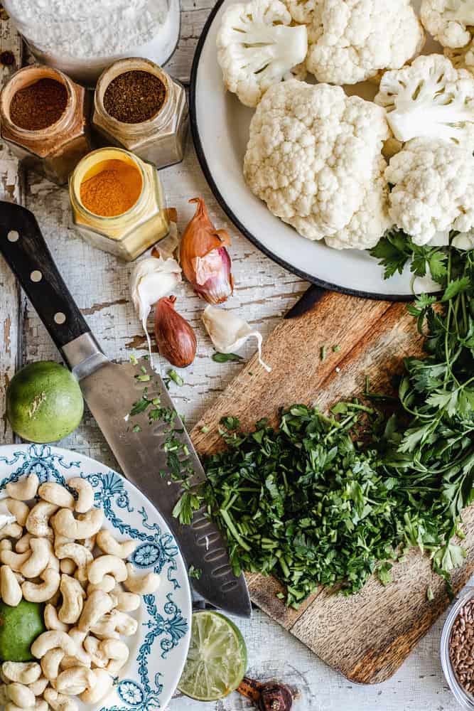 Bird's eye view of cumin cauliflower fritters and nut-yogourt sauce ingredients on a white wooden board (clockwise): cauliflower, parsley, flax seeds, cashew nuts, lime, garlic, shallots, curry powder, cumin powder, and cinnamon