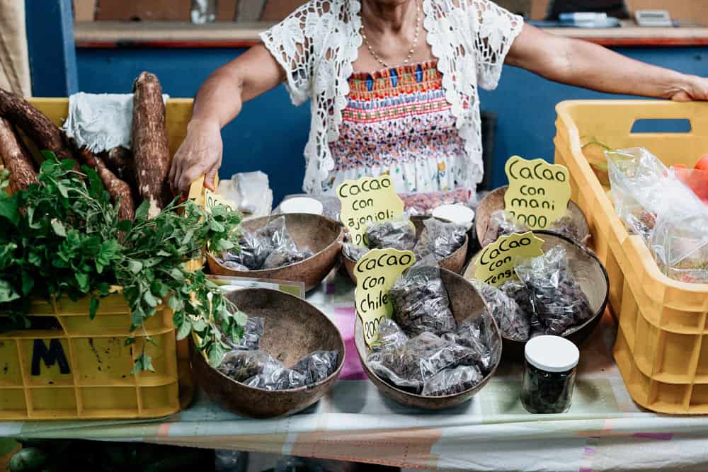 Baskets of small bags of cacao between two yellow with an old lady standing behind the stand