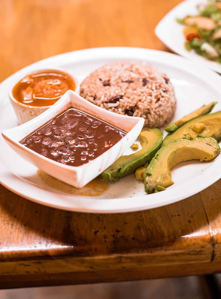 Une assiette de sauce aux haricots et de sauce tomate dans de petits bols avec du riz des Caraïbes dans une demi-boule à côté de tranches d'avocat et de haricots - Plat végétalien personnalisé chez Lidia's Place