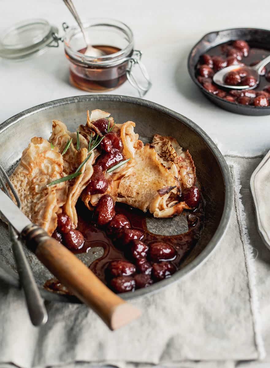 A close-up of folded pancakes in a metallic plate with strawberry sauce dripping with a small jar of maple syrup behind it and a small skillet of strawberry sauce on the side