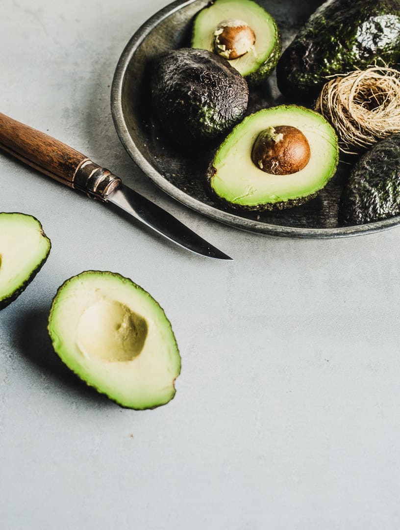 Avocado cut in half next to a metallic plate of cut and whole avocadoes