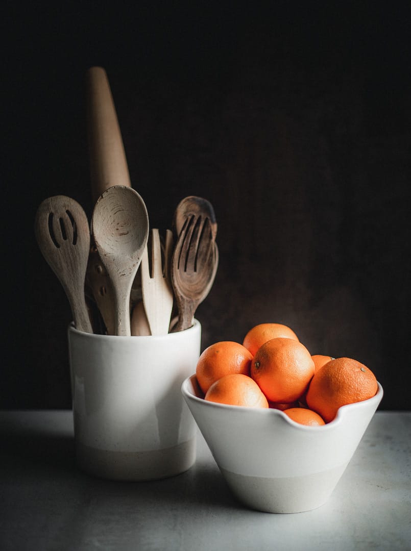 Wooden spoons and forks in a big two-toned (white and sand) ceramic canister next to a matching mixing bowl with oranges in it