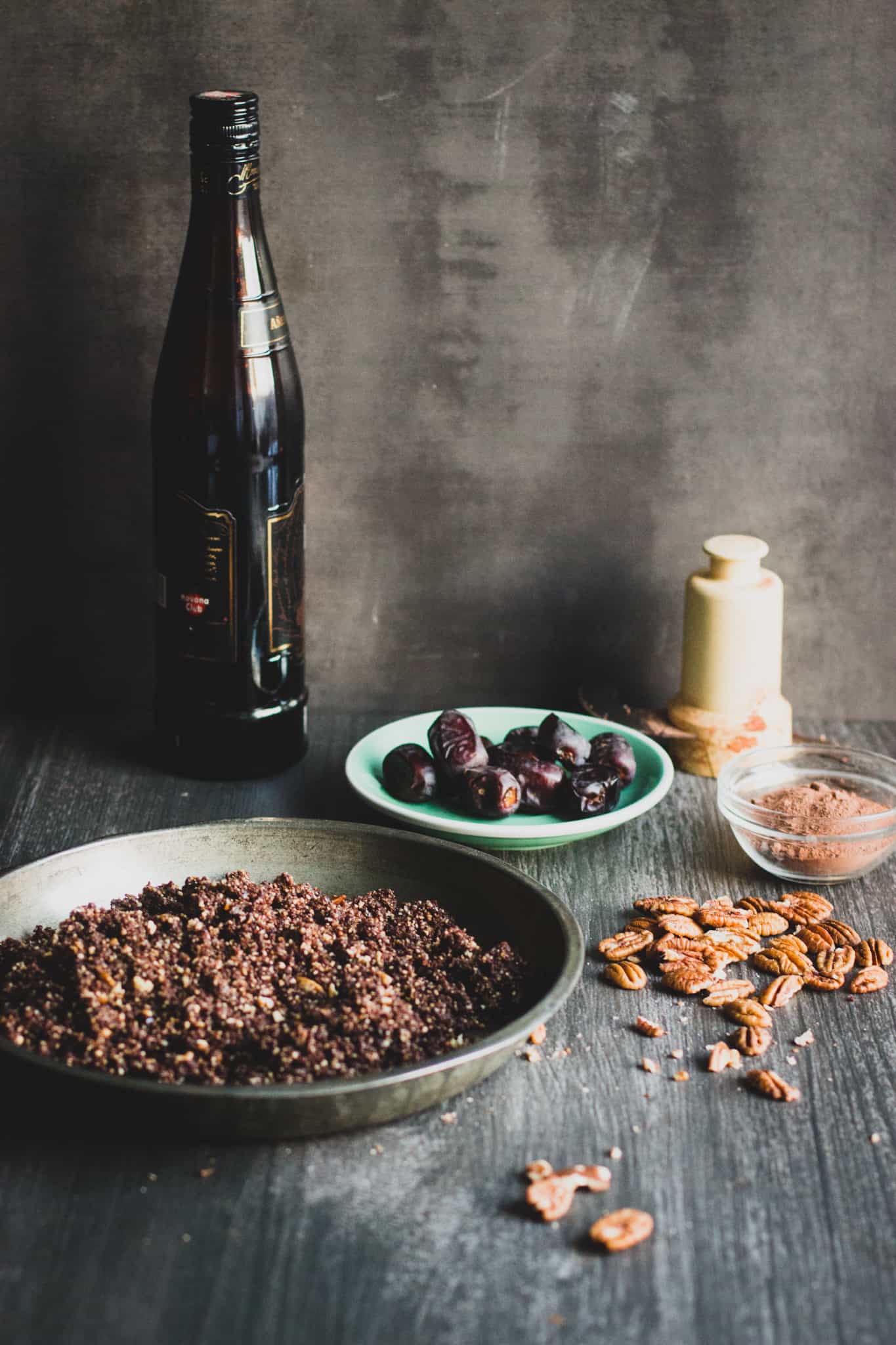 Chocolate pecan pie dough in a metallic plate in front of a bottle of rhum and next dried figs on small plate, cocoa powder in a tiny glass mixing bowl, and almonds and walnuts spread on the surface