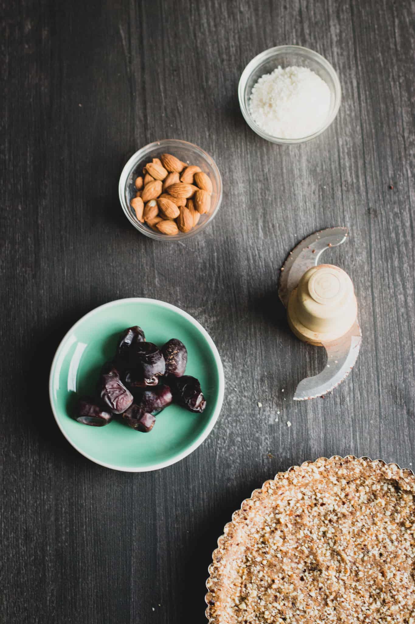 Top view of pecan pie ingredients in small mixing bowls. From top to bottom: coconut flakes, almonds, dates, and almond-walnut crust