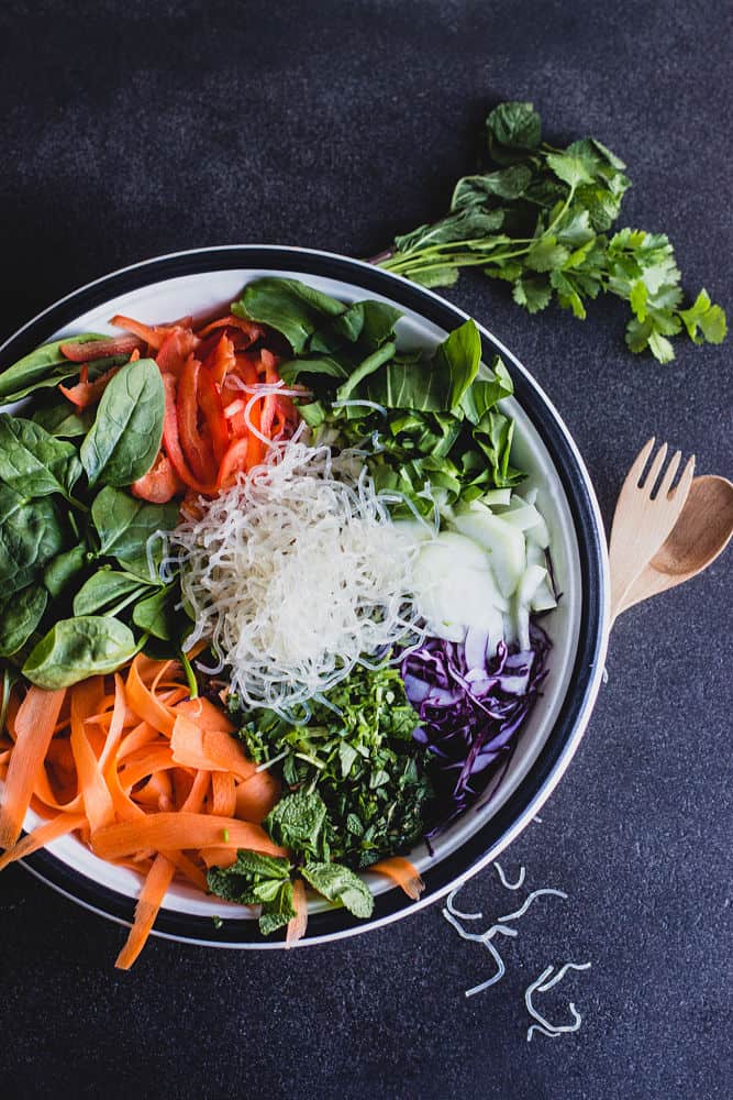 Top view of thai salad ingredients in a bowl. From left to right clockwise: spinach, red bell peppers, bok choy, red cabbage, cilantro, carrots in ribbons, and kelp noodles