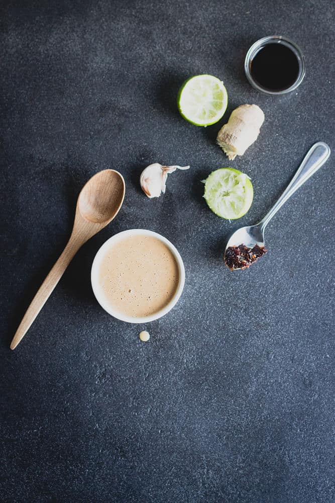 Top view of mock peanut sauce in a ramekin, garlic clove, lime cut in two, miso paste on a spoon, piece of ginger, and tamari in a tiny glass mixing bowl