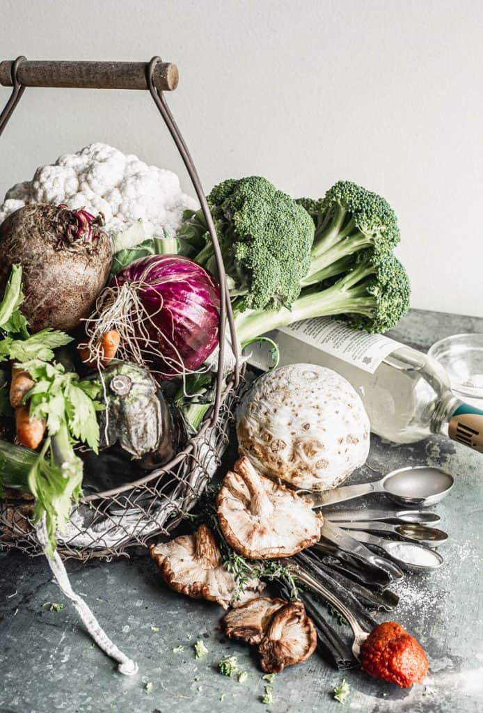 vegetables overflowing from a rustic wire basket with beside it, a bottle of wine lying down, a set of measuring spoons filled with pectin, olive oil, salt, and tomato paste, on top whit celeriac, sprigs of thyme, and shiitake mushrooms on top