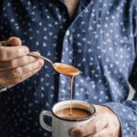 A man, dressed in a dark blue shirt, holds a spoon in his left hand and pours brown sauce into a white enamel mug that he's holding in his right hand]