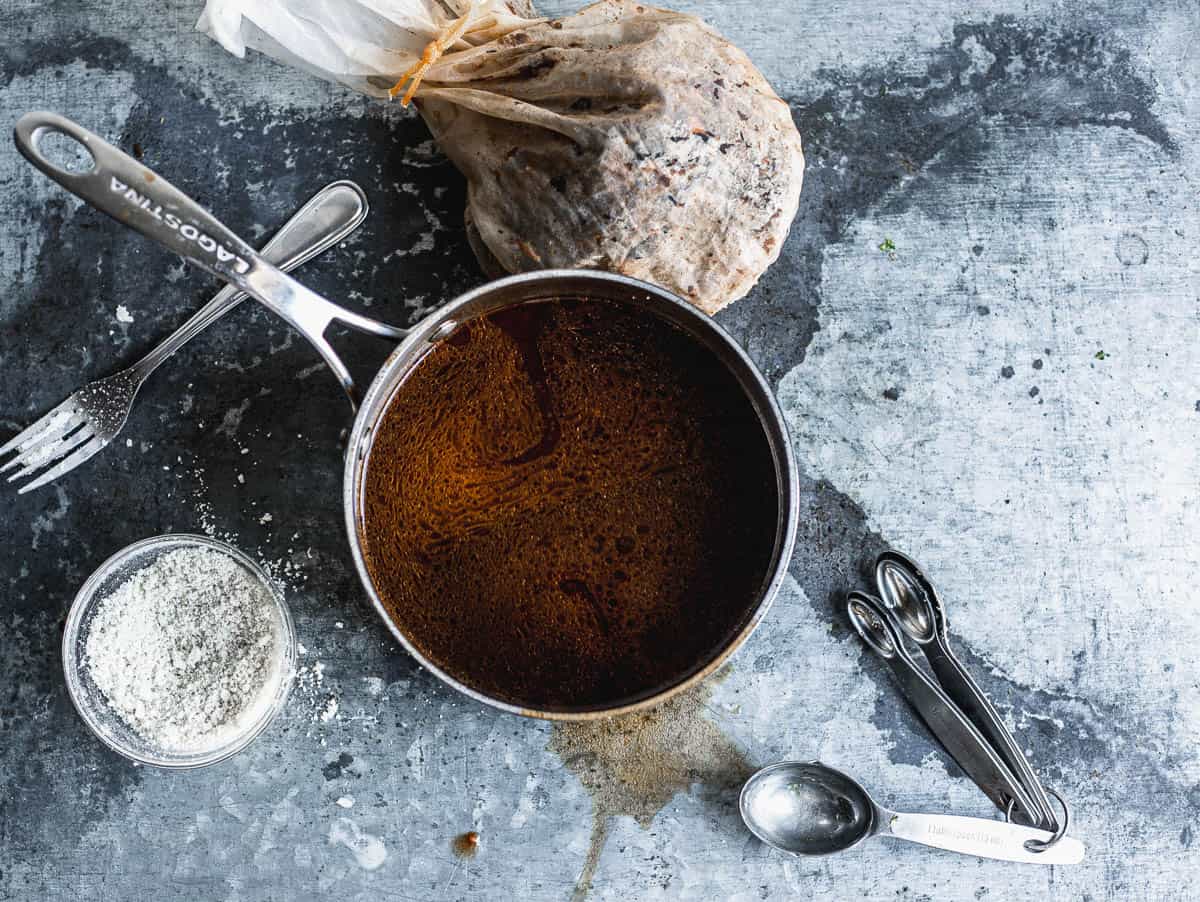 Overhead view of a small bowl of white powder next to a saucepan filled with brown aqueous sauce in a saucepan and a set of measuring spoons