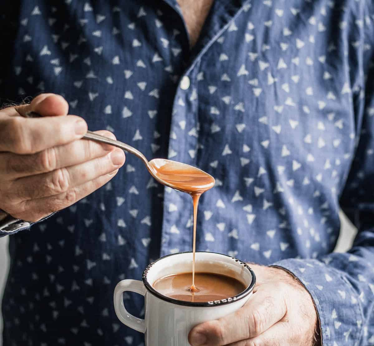 Man dressed in a printed shirt holding a spoon in his left hand and pouring brown sauce into a white enamel mug he is holding with his right hand