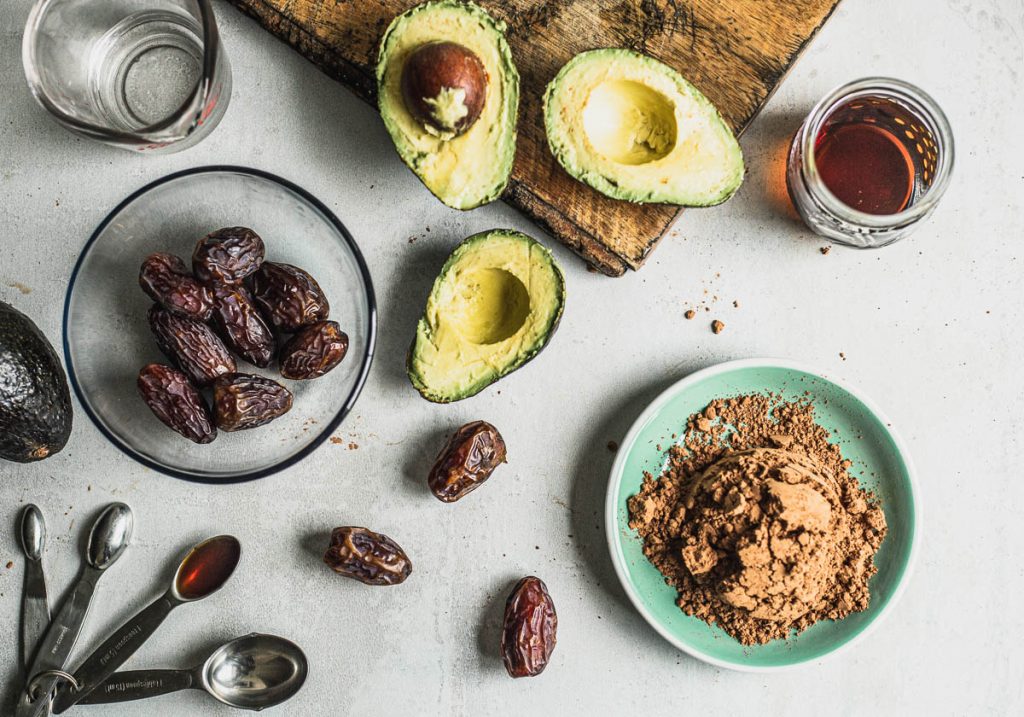 Top view of vegan chocolate mousse ingredients (clockwise): Medjool dates in a glass bowl, a pyrex measuring cup with water, avocados cut in half, maple syrup in a small jar, cocoa powder in a small plate, scattered Medjool plates, and vanilla extract in a set of measuring spoons.