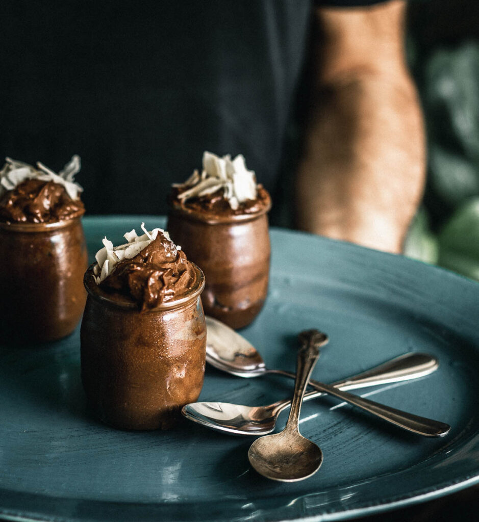 Chocolate mousse in three small jars with large coconut flakes on top on a plate being held by a man in the background