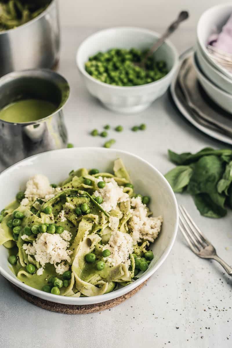 Pappardelle-style pasta with peas and ricotta in a rustic bowl, gravy boat with green sauce, peas in a bowl, stacked plates, a bunch of basil, ricotta in a ramekin