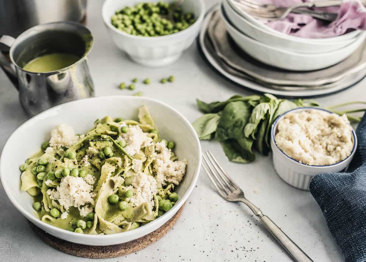 Pappardelle-style pasta with peas and ricotta in a rustic bowl, gravy boat with green sauce, peas in a bowl, stacked plates, a bunch of basil, ricotta in a ramekin