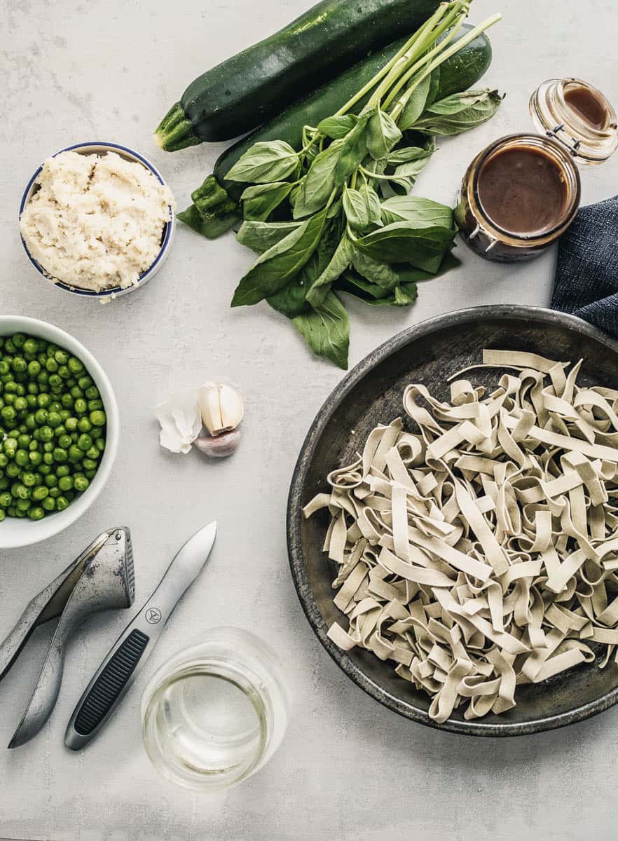 Ingredients spread out on a white plate (clockwise): overhead view of peas in a bowl, ricotta in a ramekin, a bunch of basil on top of the zucchini, a brown sauce in a mini Mason jar, ribbons of dry pasta in a metal plate, cloves of garlic.
