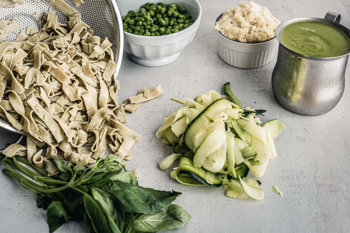 Pappardelle-style pasta overflowing with a sieve with on their left a bunch of basil and on their right, peas in a bowl, ricotta in a ramekin, a green sauce in a gravy boat and zucchini finely cut into ribbons.