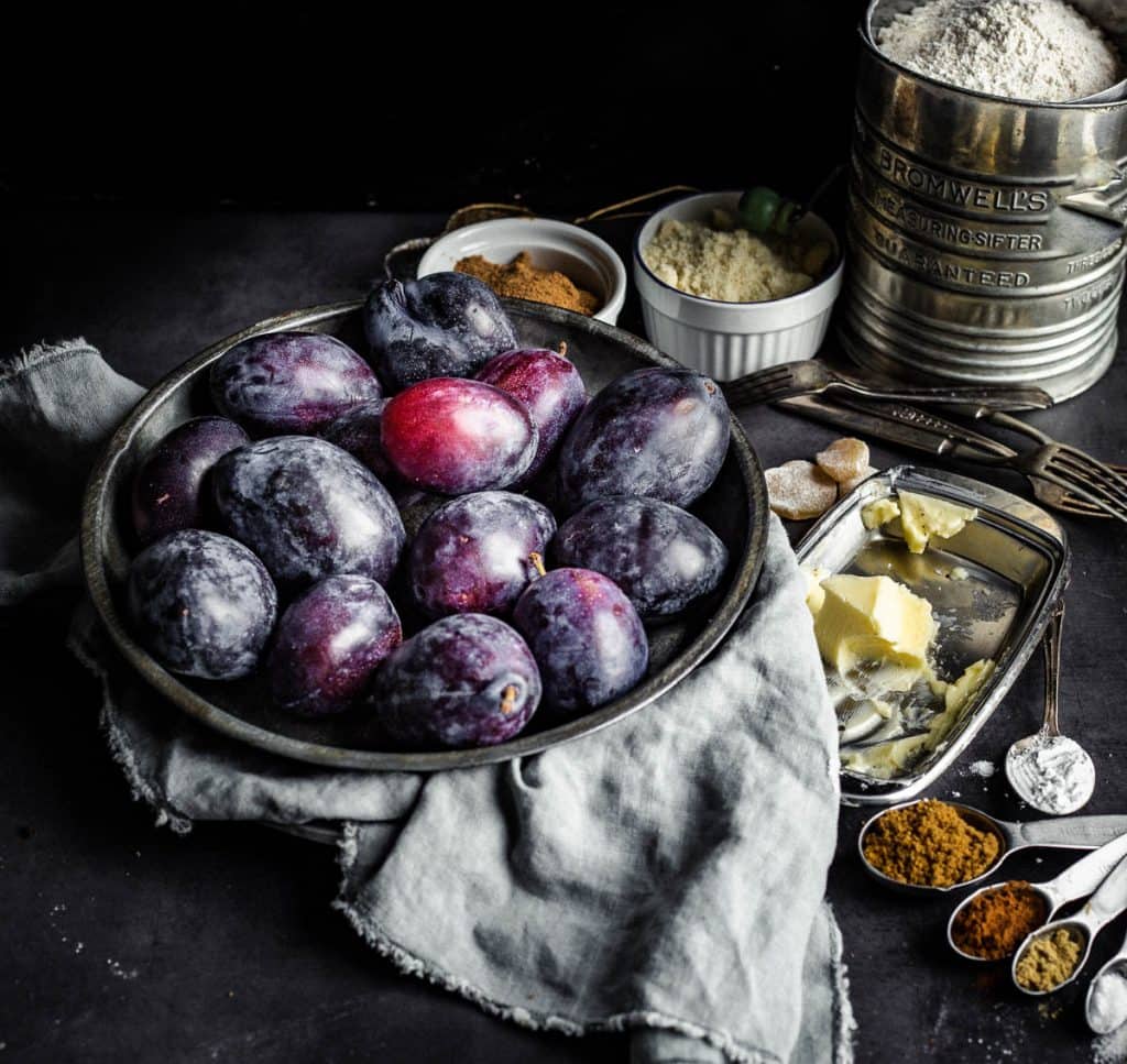 Oblong plums in a metallic plate surrounded by different types of flours in bowls, butter on a butter dish, and spices in measuring spoons