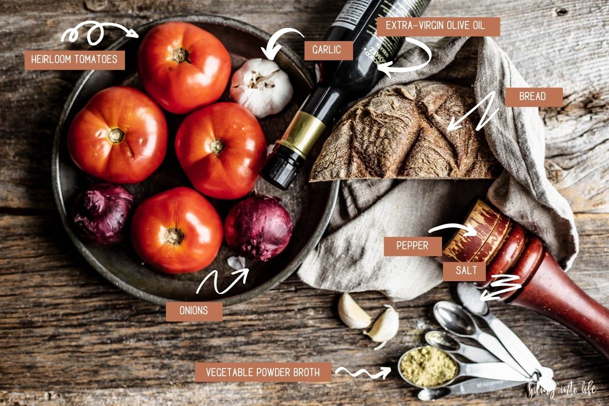 Overhead view of the ingredients for the pappa al pomodoro (clockwise): tomatoes, garlic, red onions in a metal bowl with to their right a bottle of olive oil, a half of a loaf of bread , a pepper pot and a set of measuring spoons with powdered vegetable broth and salt