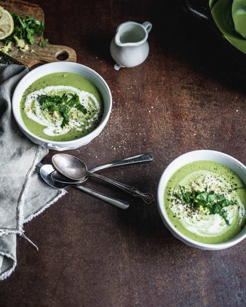 Two bowls of green soup garnished with white cream in a spiral, parsley and lemon zest and placed diagonally