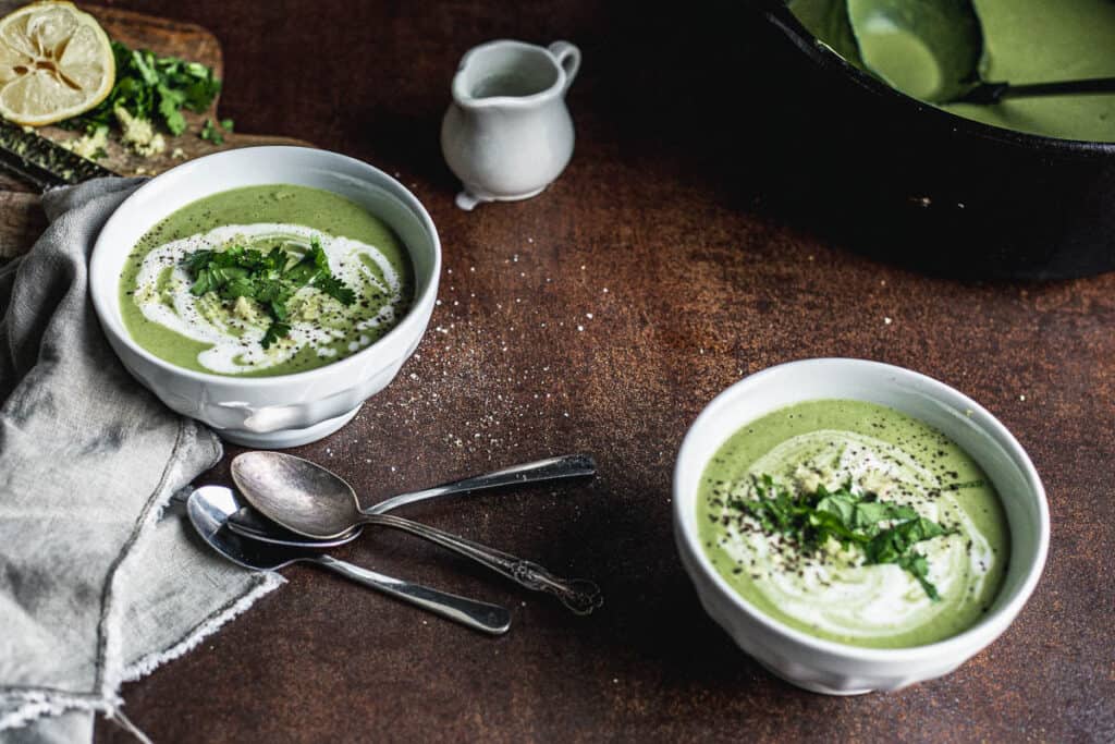 Two bowls of green soup garnished with white cream in a spiral, parsley and lemon zest and placed diagonally