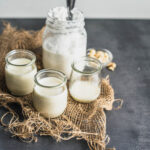 Homemade vegan half-and-half in three small glass jars and in front of a small mason jar of coconut cream, all on a piece of jute