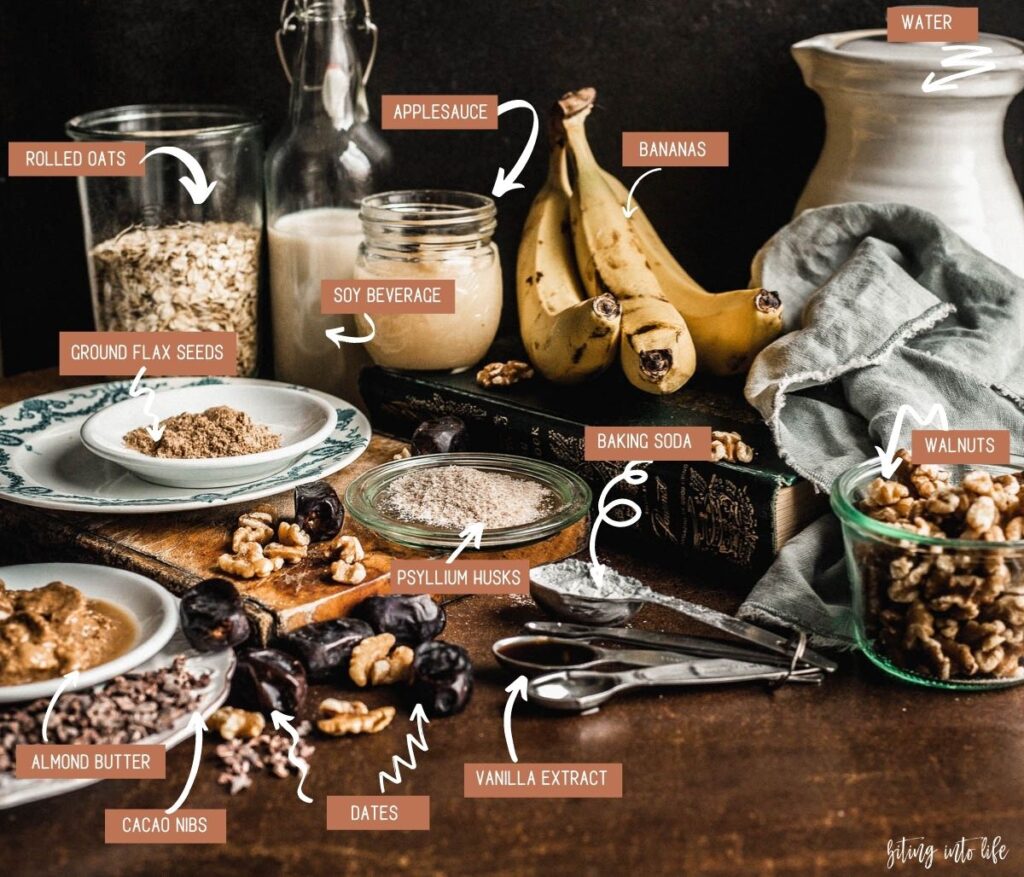Banana bread Ingredients (clockwise): cocoa nibs and almond butter on small plates, ground flax seeds on a small plate, rolled oat in a glass jar, soy beverage in a small bottle, applesauce in a small glass jar and three bananas placed on a thick book, walnuts in a jar, baking powder and vanilla extract in a set of spoons, psyllium husks on a glass lid, and dates and scattered walnuts