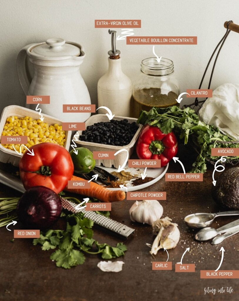 Black Bean Soup Ingredients (clockwise): corn kernels in a small cardboard container, black beans in a small cardboard container, red bell pepper, extra-virgin olive oil in its oil dish, vegetable broth concentrate in a Mason jar, bunch of cilantro in a wire basket, avocado, grains of salt and pepper scattered on the surface, garlic cloves, chili powder, ground cumin, chipotle, kombu, lime, tomato and Red onion