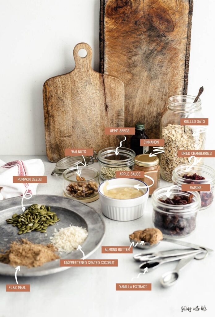 Vegan granola ingredients (clockwise): metallic dish containing pumpkin seeds, unsweetened grated coconut flakes next to lots of small jars of walnuts, hemp, cinnamon powder, rolled oats, cranberries, applesauce, and a set of measuring spoons containing almond butter and vanilla extract