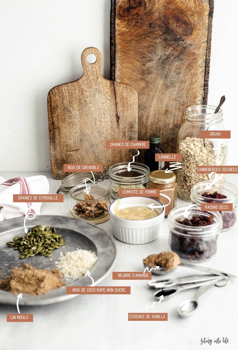 Vegan granola ingredients (clockwise): metallic dish containing pumpkin seeds, unsweetened grated coconut flakes next to lots of small jars of walnuts, hemp, cinnamon powder, rolled oats, cranberries, applesauce, and a set of measuring spoons containing almond butter and vanilla extract