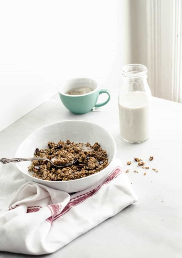 A bowl of granola on a white counter accompanied by a small bottle of soy beverage and a cup of tea