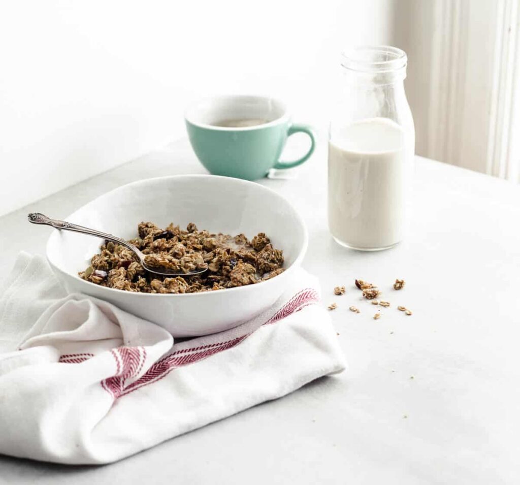 A bowl of granola on a white counter accompanied by a small bottle of soy beverage and a cup of tea