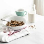 A bowl of granola on a white counter accompanied by a small bottle of soy beverage and a cup of tea