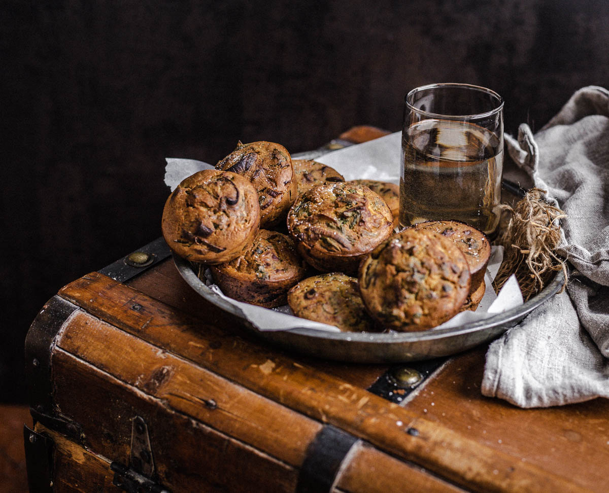 A batch of savory muffins in a metal plate resting on a wooden chest and torn muffin on a narrow cutting board.