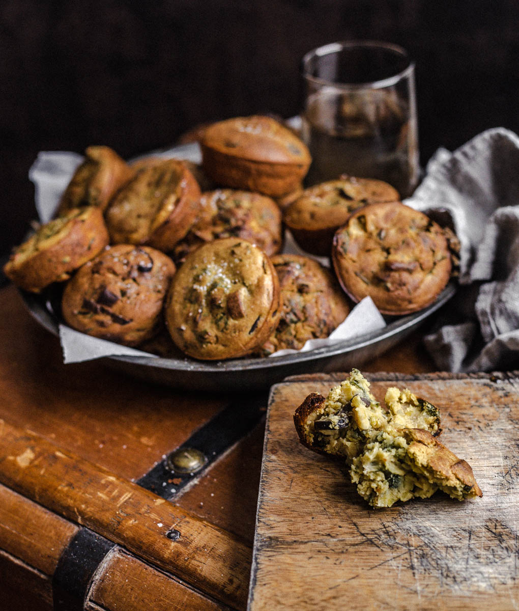 A batch of savory muffins in a metal plate resting on a wooden chest and torn muffin on a narrow cutting board.