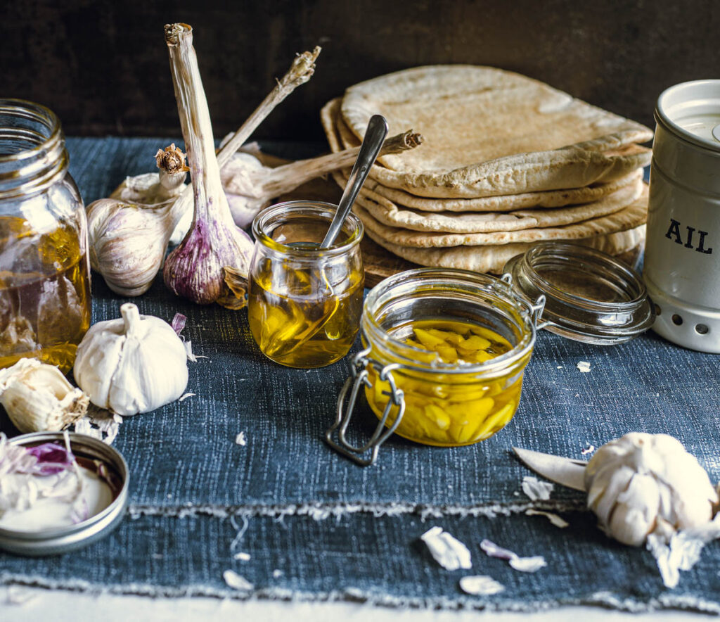 Three small glass jars of garlic-infused oil, surrounded by decorative garlic bulbs, one of which contains garlic cloves cut in half and the others have only the oil