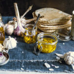 Three small glass jars of garlic-infused oil, surrounded by decorative garlic bulbs, one of which contains garlic cloves cut in half and the others have only the oil